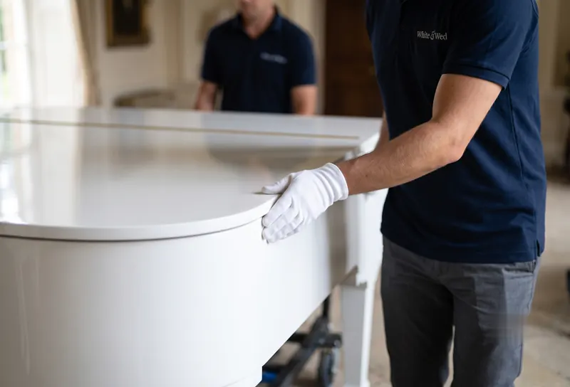 White & Wed crew member in white gloves steadying the white grand piano during delivery at a wedding venue